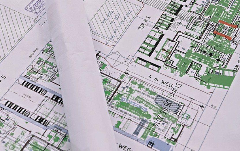 Farbige Baupläne mit Grundriss, Wegführung und Infrastrukturdetails, ausgebreitet auf Tisch, mit gerolltem Papier im Vordergrund